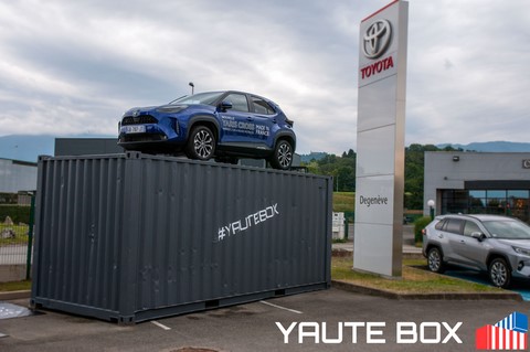 container avec une toyota yaris sur le toit devant le panneau de toyota blanc ecriture rouge